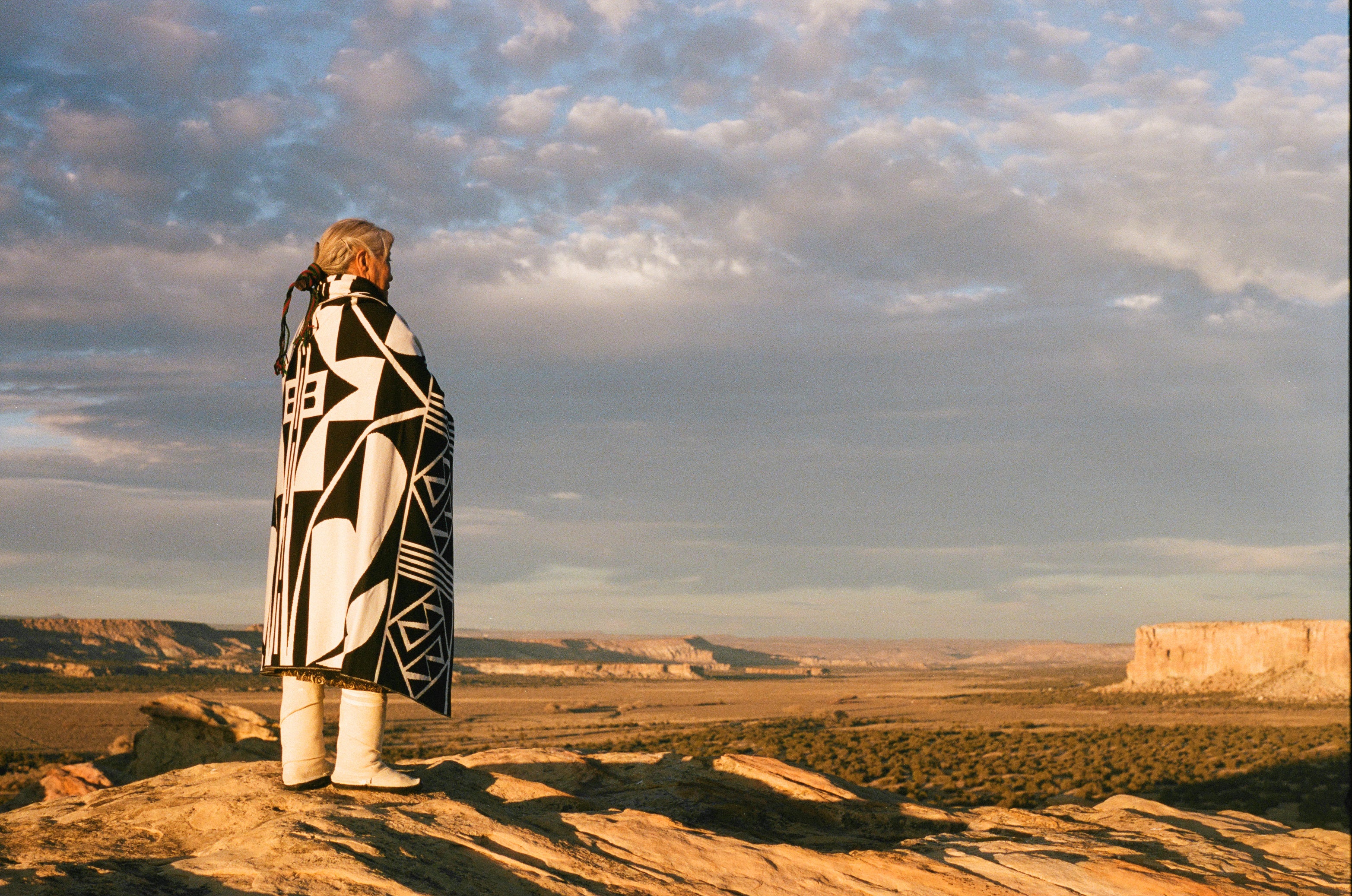 Person wrapped in a patterned blanket standing on a rocky outcrop with a vast landscape and cloudy sky.